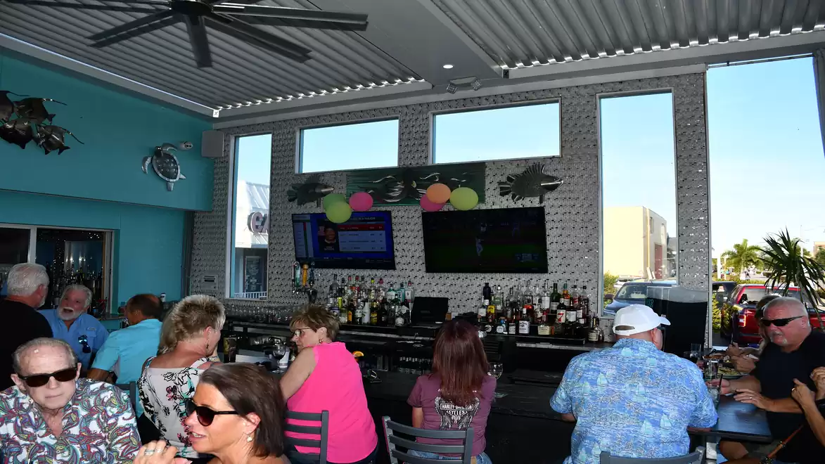 Guests in a bar enjoying their drinks, illuminated by bright daylight with large windows and several TVs on the wall.