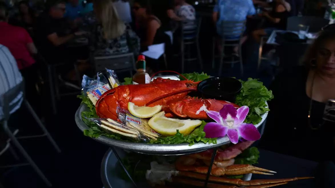 A plate with a large red lobster, surrounded by lemon slices, kale leaves, and a purple orchid flower in a busy restaurant.