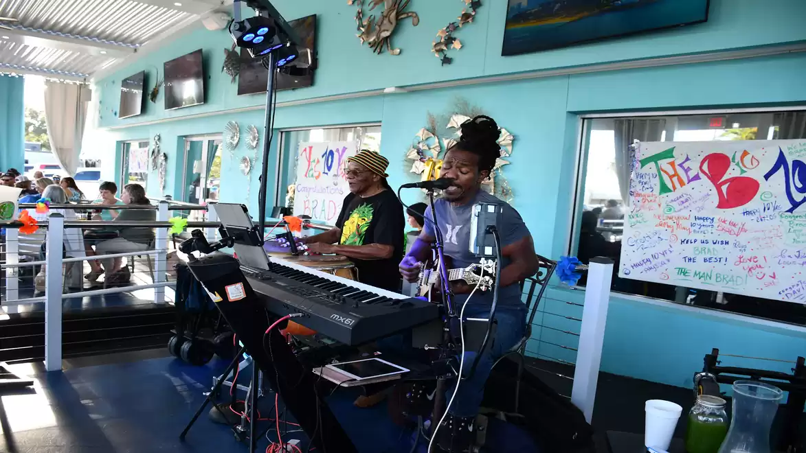 Two musicians perform live music in a bright, modern restaurant with a sea view. One musician plays the keyboard and sings, while the other plays drums.