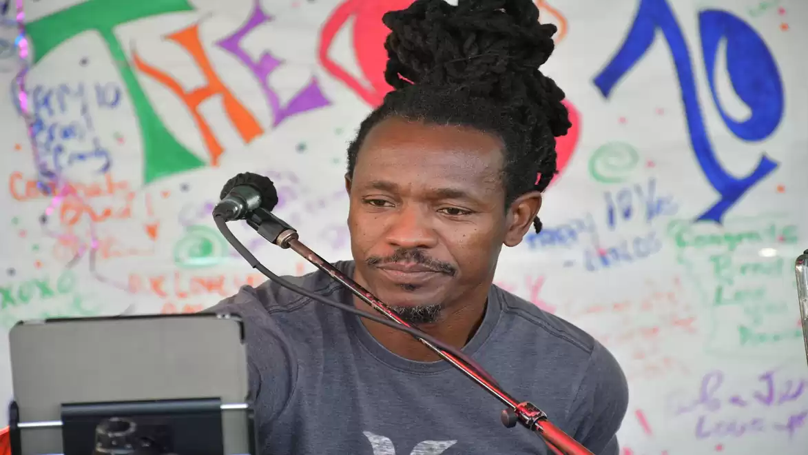 A man with dreadlocks speaks into a microphone in front of a colorfully decorated wall.