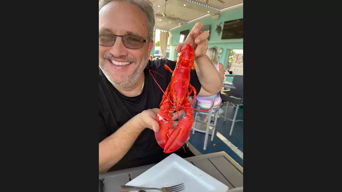 A man sitting in a restaurant holds a large red lobster towards the camera while smiling.