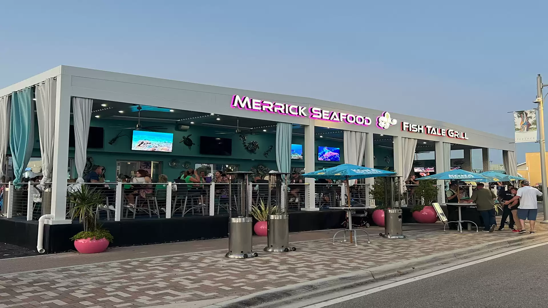 A modern restaurant named Fish Tale Grill during daylight, featuring blue and white curtains and guests seated outside. Bright neon lighting and several screens are visible.