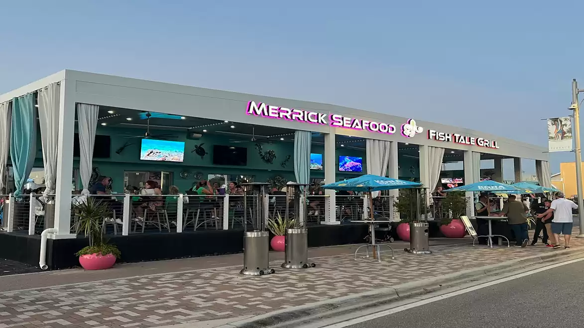 A modern restaurant named Fish Tale Grill during daylight, featuring blue and white curtains and guests seated outside. Bright neon lighting and several screens are visible.