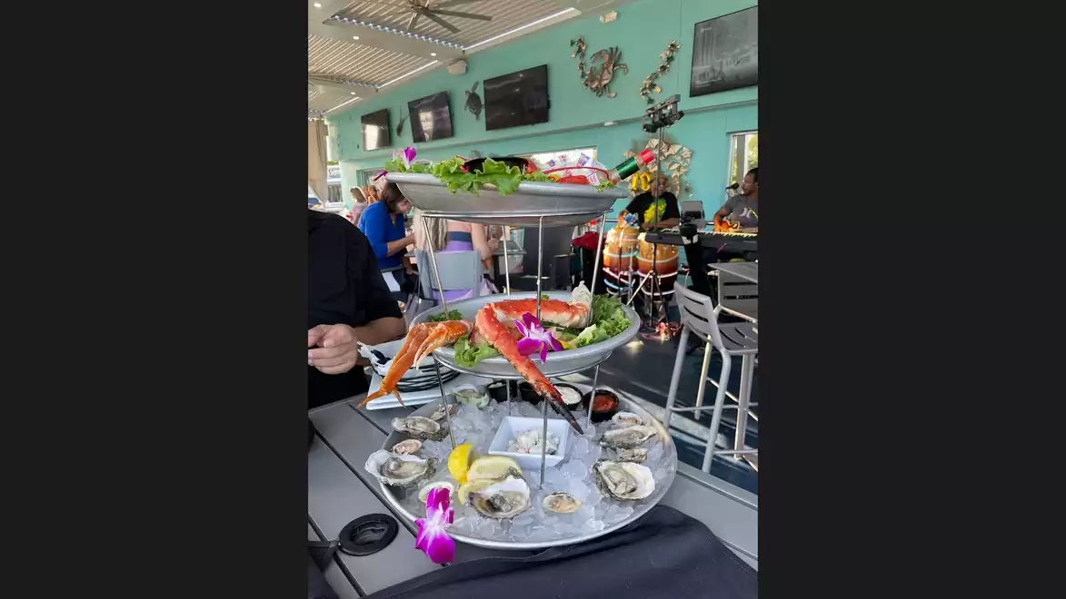 A multi-tiered serving platter with seafood, including crab legs and oysters, decorated with edible flowers. Restaurant guests are visible in the background.