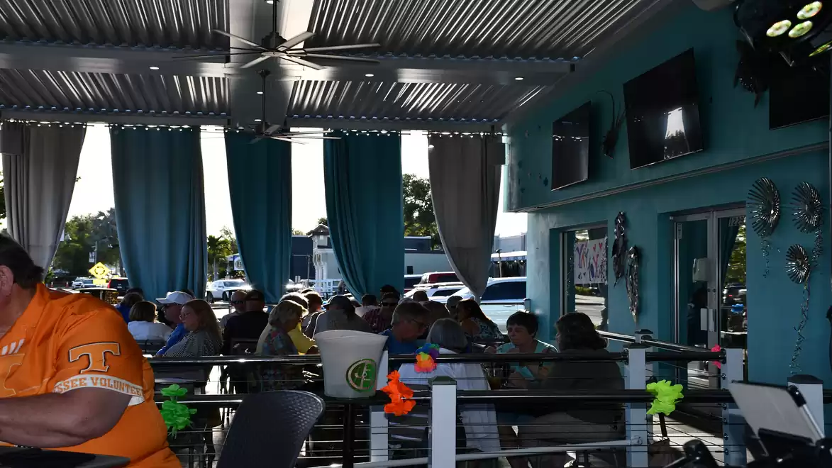 A group of people sitting in a restaurant with an open roof and blue curtains during daylight. The walls are turquoise and there are several televisions on the wall.