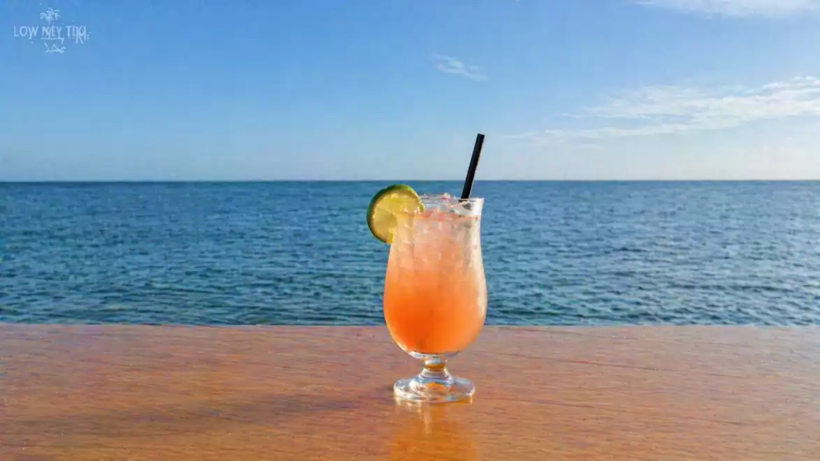 A refreshing cocktail with a lime slice on the glass sits on a wooden table overlooking a calm sea under a clear sky.