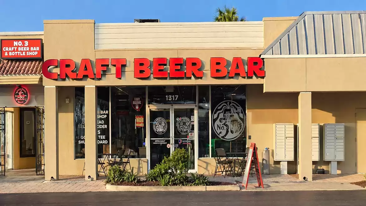 Facade of a craft beer bar with a red sign and glass front, patio with seating.