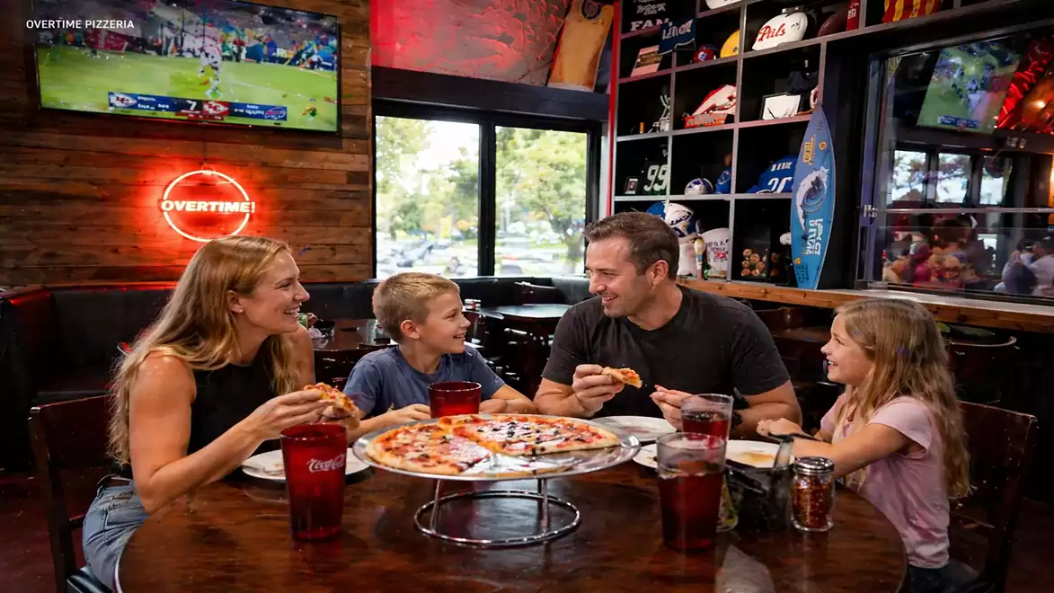 A family is sitting together in a pizzeria enjoying pizza. Sports items and a TV are visible in the background.