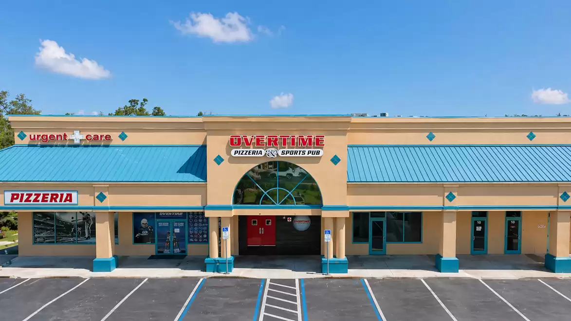 Exterior view of a beige shopping center with a turquoise roof. In the middle is a store with the sign 'OVERTIME PIZZERIA SPORTS PUB'. On the left is a sign for a pizzeria, and on the right is an urgent care sign.