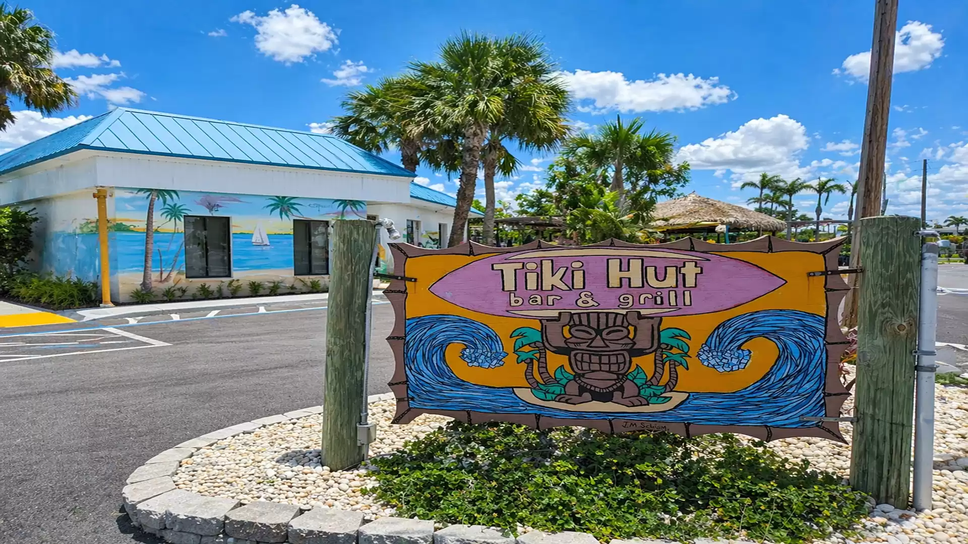 A colorful sign reading 'Tiki Hut bar & grill' stands in front of a building with blue and yellow murals, surrounded by palm trees on a sunny day.