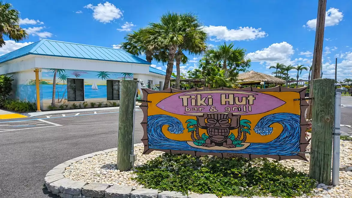A colorful sign reading 'Tiki Hut bar & grill' stands in front of a building with blue and yellow murals, surrounded by palm trees on a sunny day.