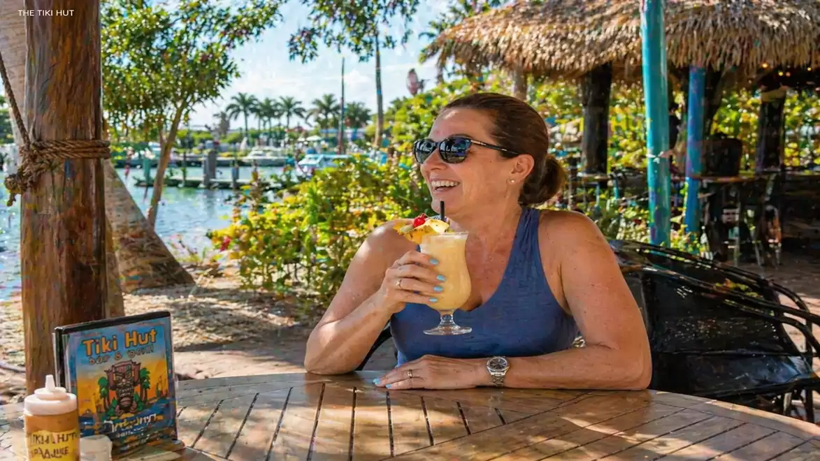 Woman in a blue tank top enjoys a tropical drink at a beach hut by the water with palm trees in the background.