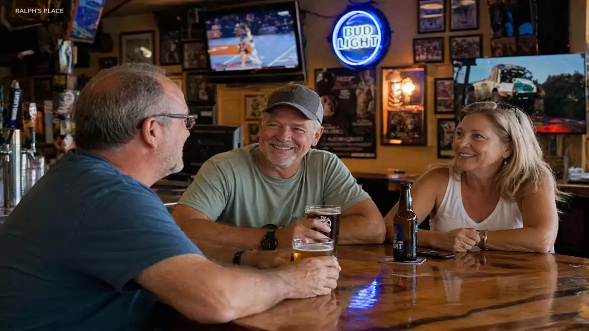 Three people sitting at a bar with drinks, surrounded by pictures and a TV in the background.