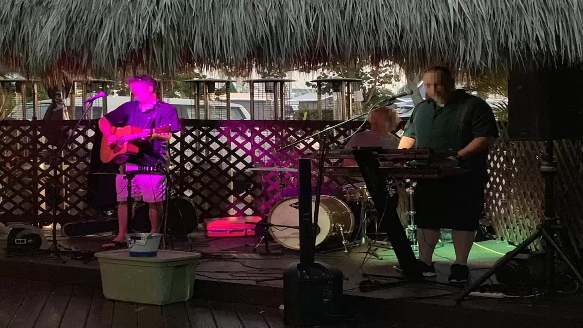 A band performs on a stage with a thatched roof. A guitarist, a drummer, and a keyboard player are visible.