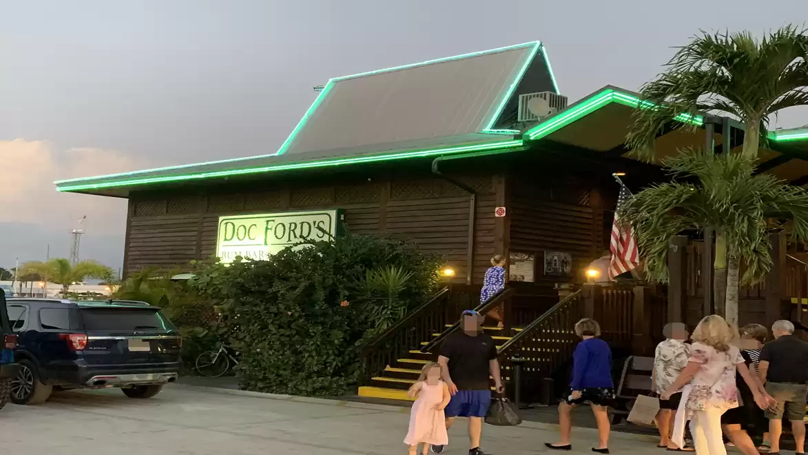 A restaurant with a glowing green roof and a group of people entering the entrance. A palm tree stands next to it.
