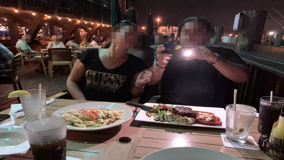Two people enjoying a dinner on a terrace with string lights in the background. On the table are a plate of pasta, a steak dish, and drinks.