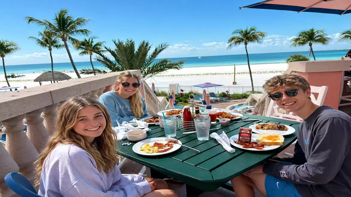 Three young adults happily dining on a terrace overlooking a sunny beach.