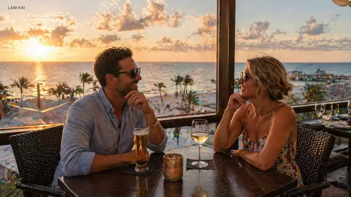 A couple sits at a table in a restaurant overlooking the sunset over the sea. They enjoy drinks and engage in conversation. Palm trees and the beach are visible in the background.