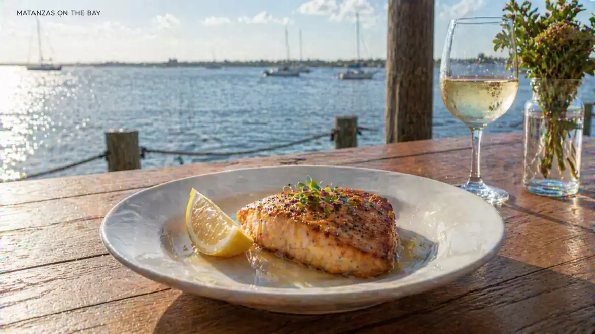 A grilled fish dish with a lemon wedge on a white plate, served on a wooden table with a wine glass, featuring a picturesque view of the water with sailboats in the background.