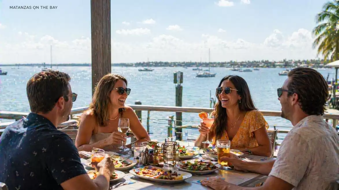 Four people dining and drinking at a table overlooking the water in sunshine.