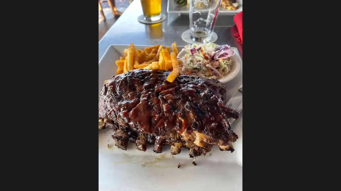 A plate of juicy BBQ ribs served with French fries and coleslaw on a restaurant table with a glass of beer in the background.