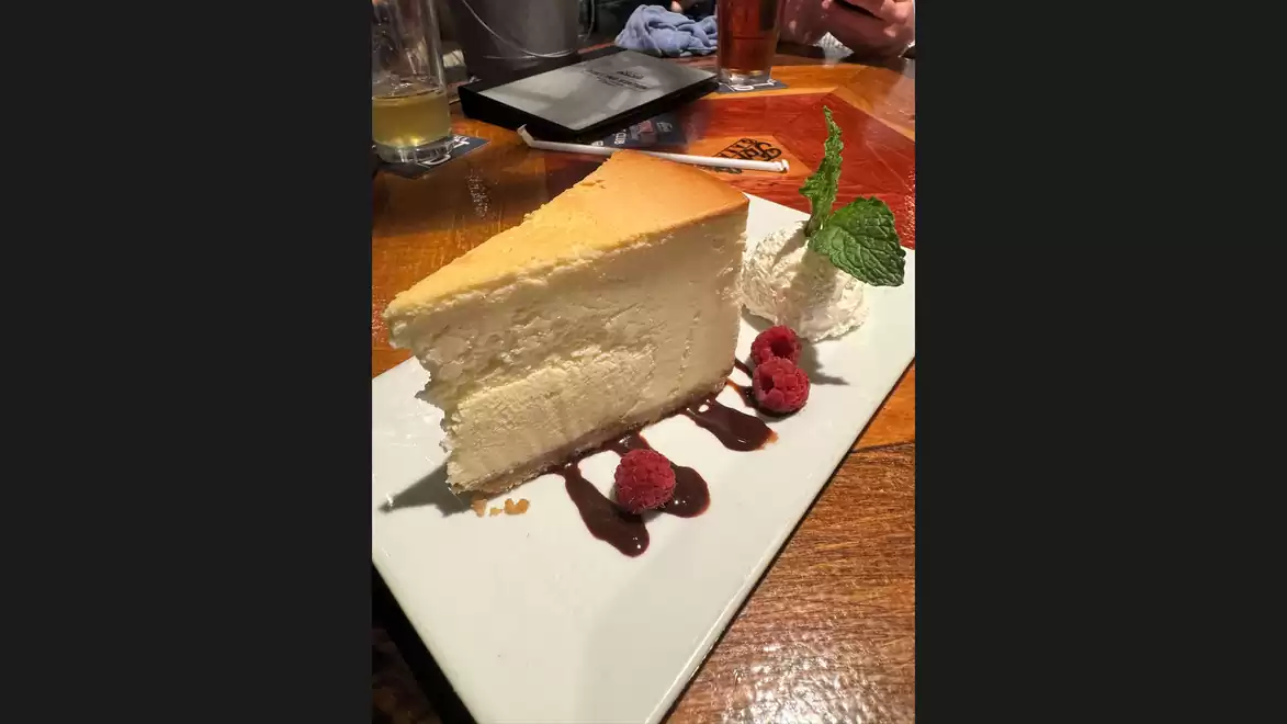 A slice of cheesecake with raspberries and cream on a white plate, decorated with mint leaves.