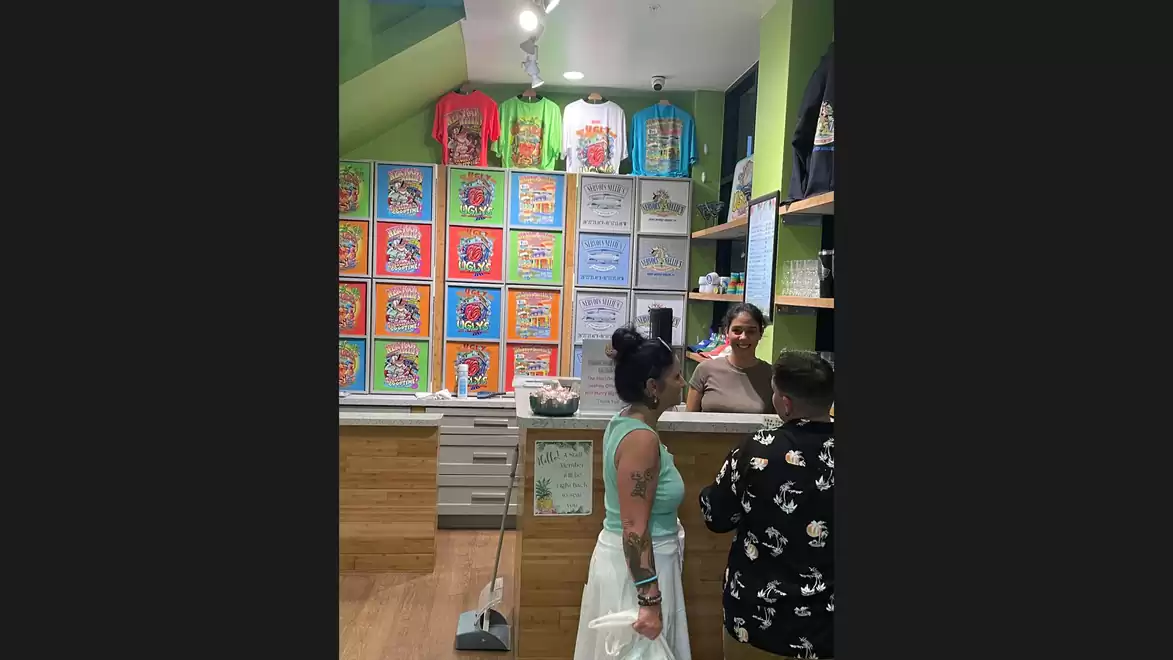 A store with colorful T-shirts and cartoon designs on the wall, with a salesperson behind the counter. Two female customers stand at the checkout.