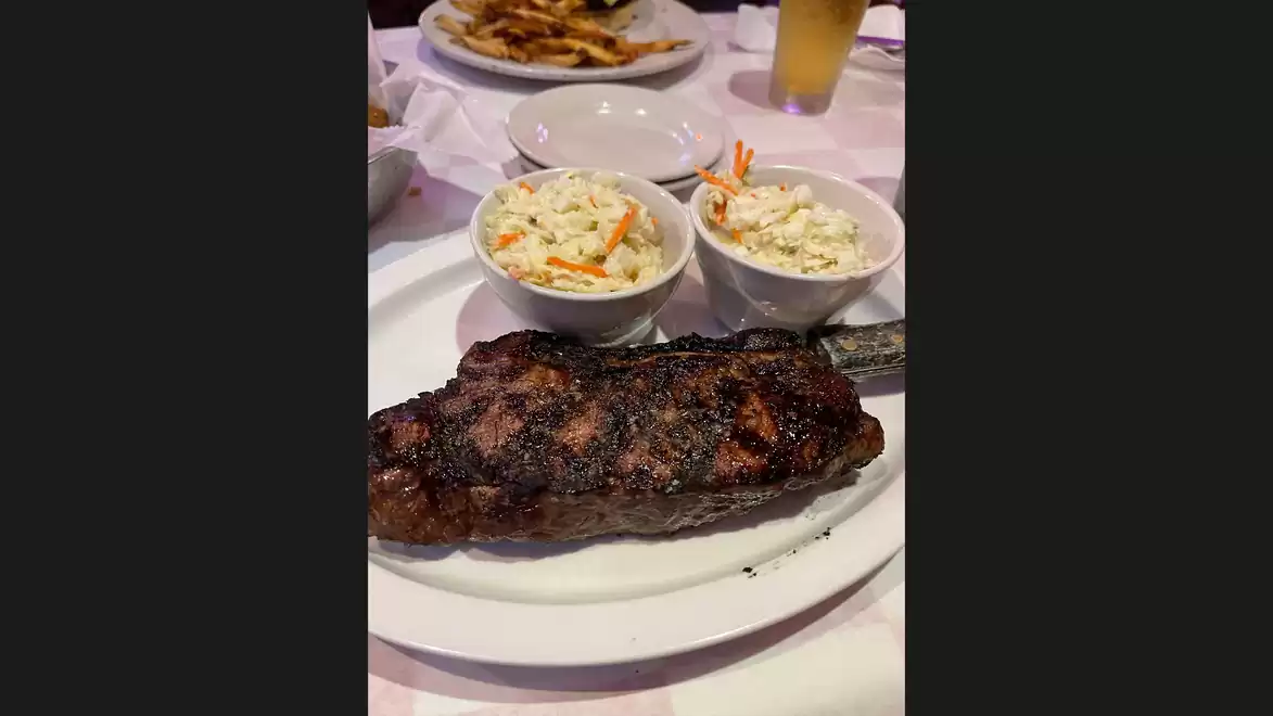 Grilled steak with two sides of coleslaw and a drink in the background on a restaurant table.