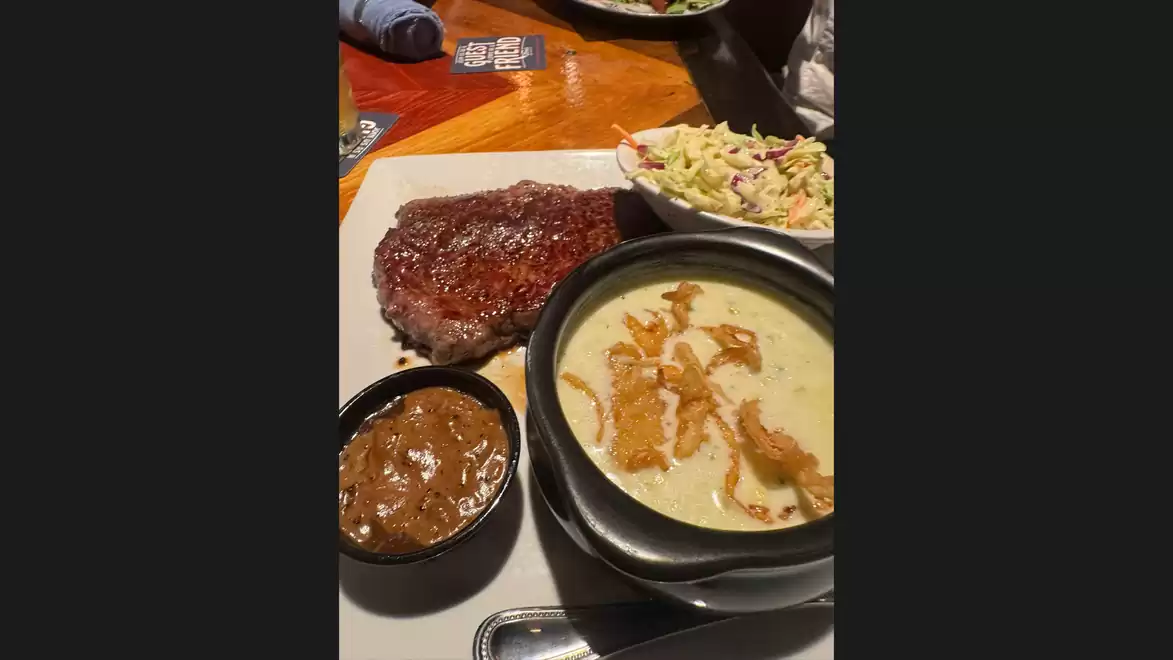 A plate featuring a juicy steak, a bowl of creamy soup, a small bowl of sauce, and a serving of coleslaw, served on a wooden table.
