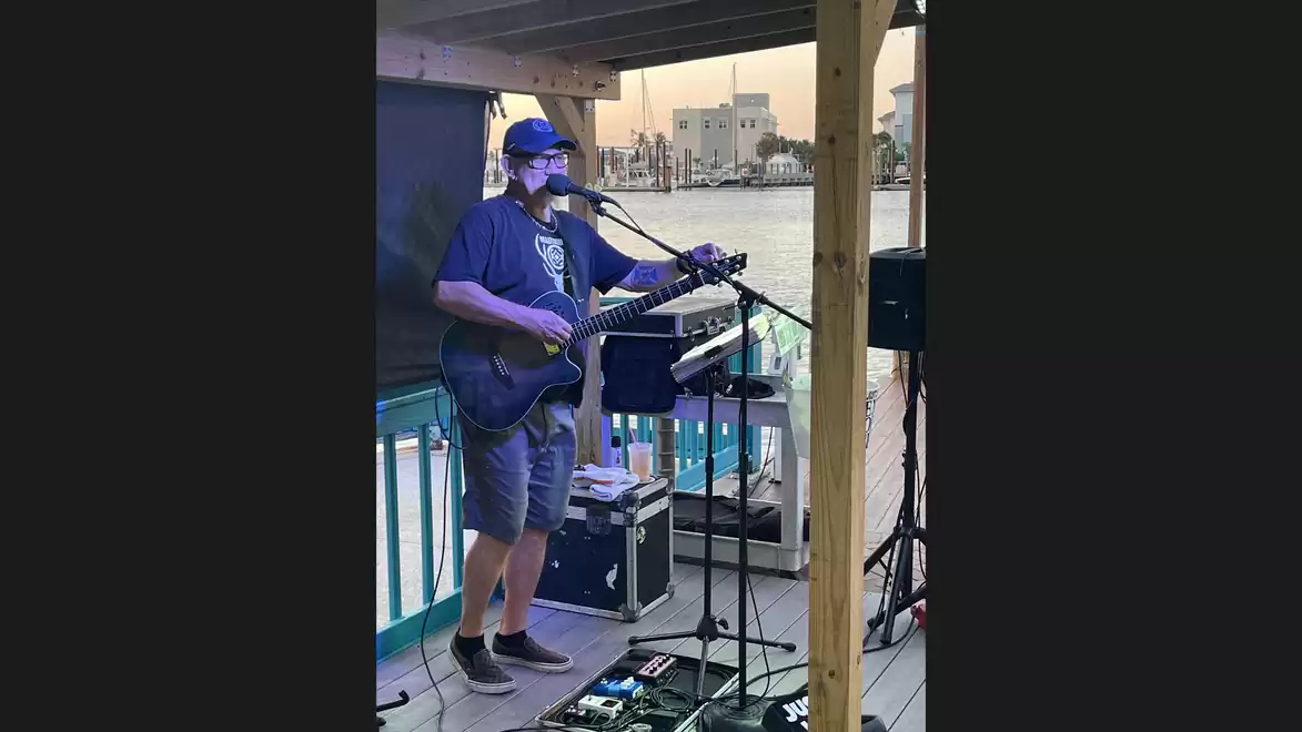 A musician plays guitar and sings on a deck overlooking a body of water in the background.