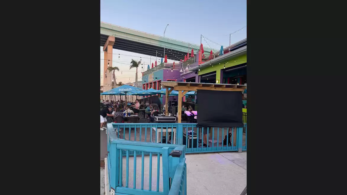 Outdoor area of a restaurant with a blue fence and tables under umbrellas, covered by a bridge in clear weather.