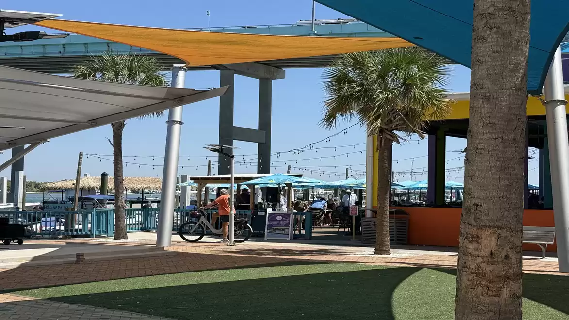 A sunny outdoor area with palm trees, colorful canopies, and a bustling café in the background.