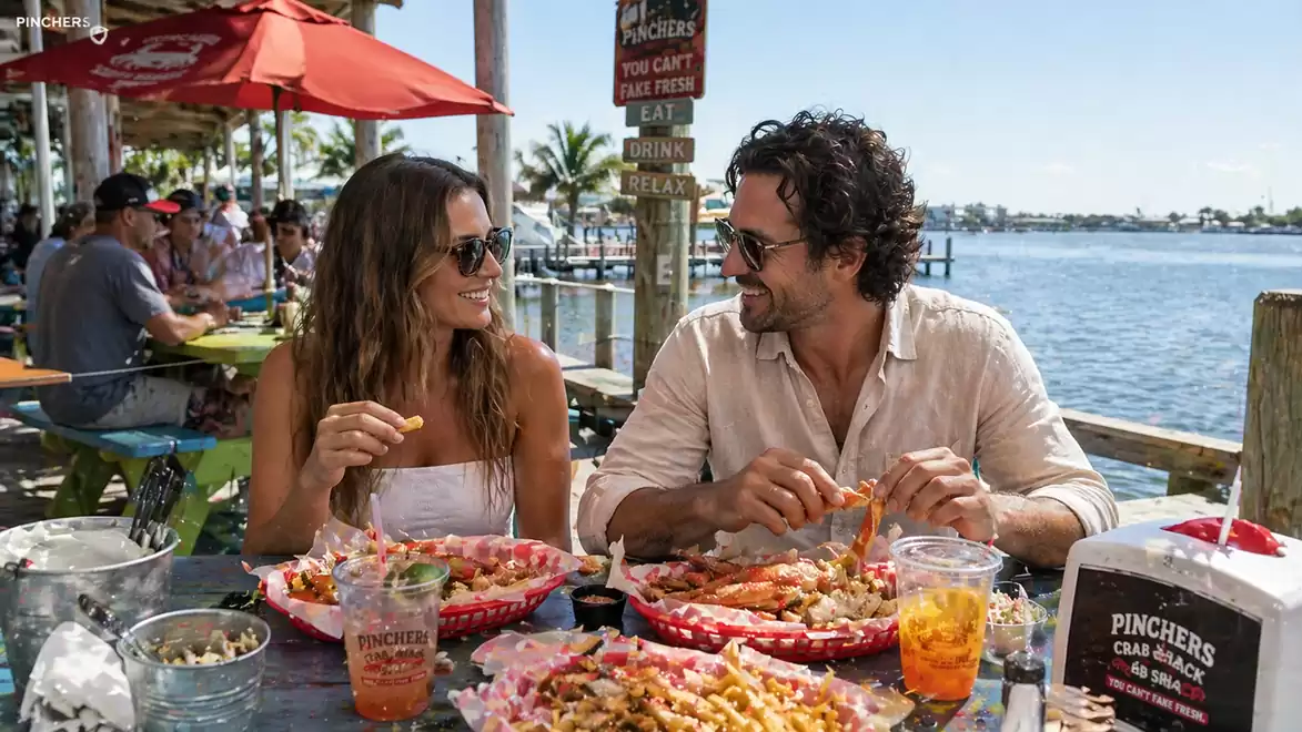 A couple sits at a table by the water enjoying a seafood meal in sunny weather at a waterfront restaurant.