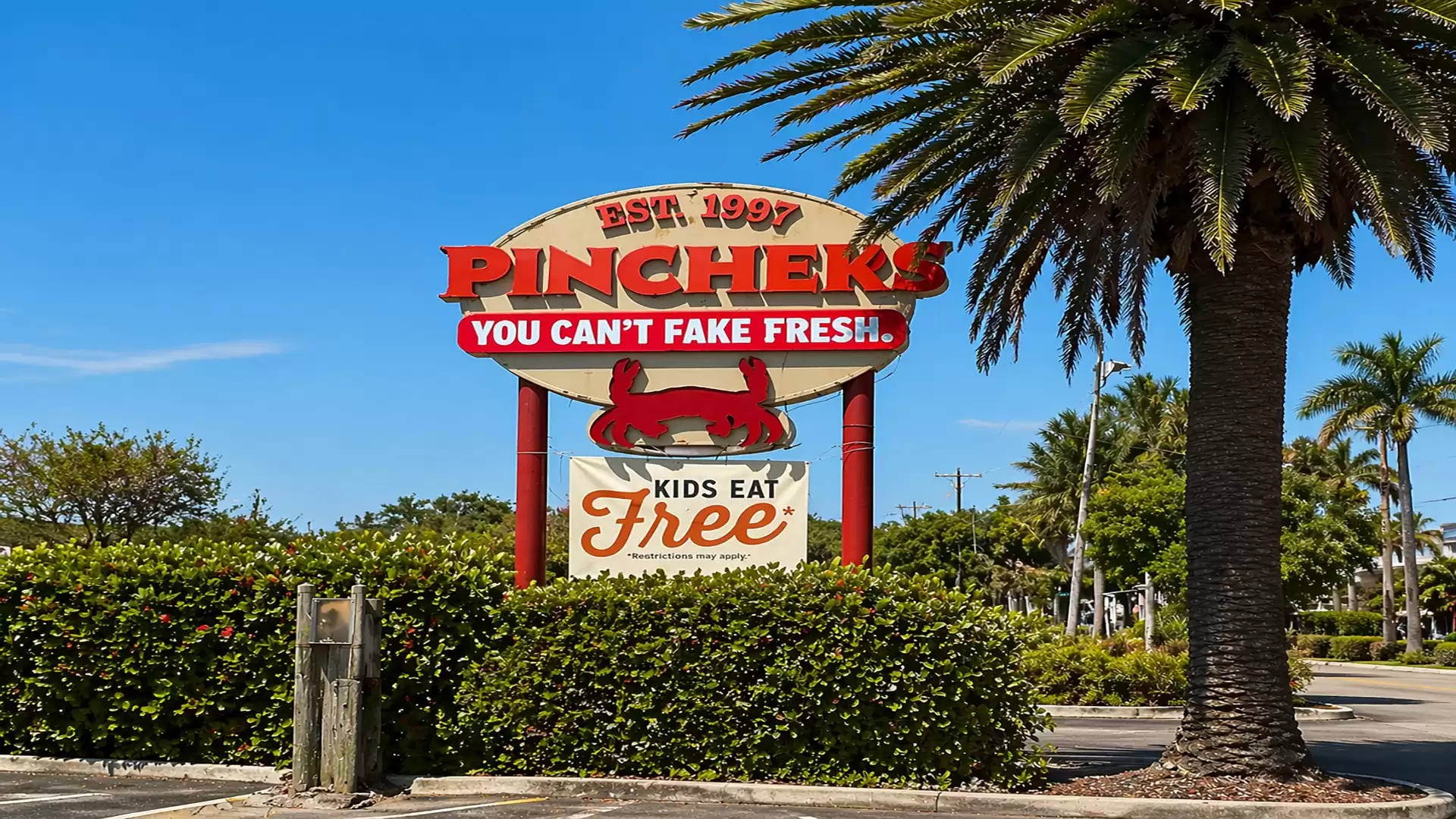 A sign for the restaurant Pinchers with the text 'You can't fake fresh' and 'Kids eat free'. Palm trees and blue sky in the background.