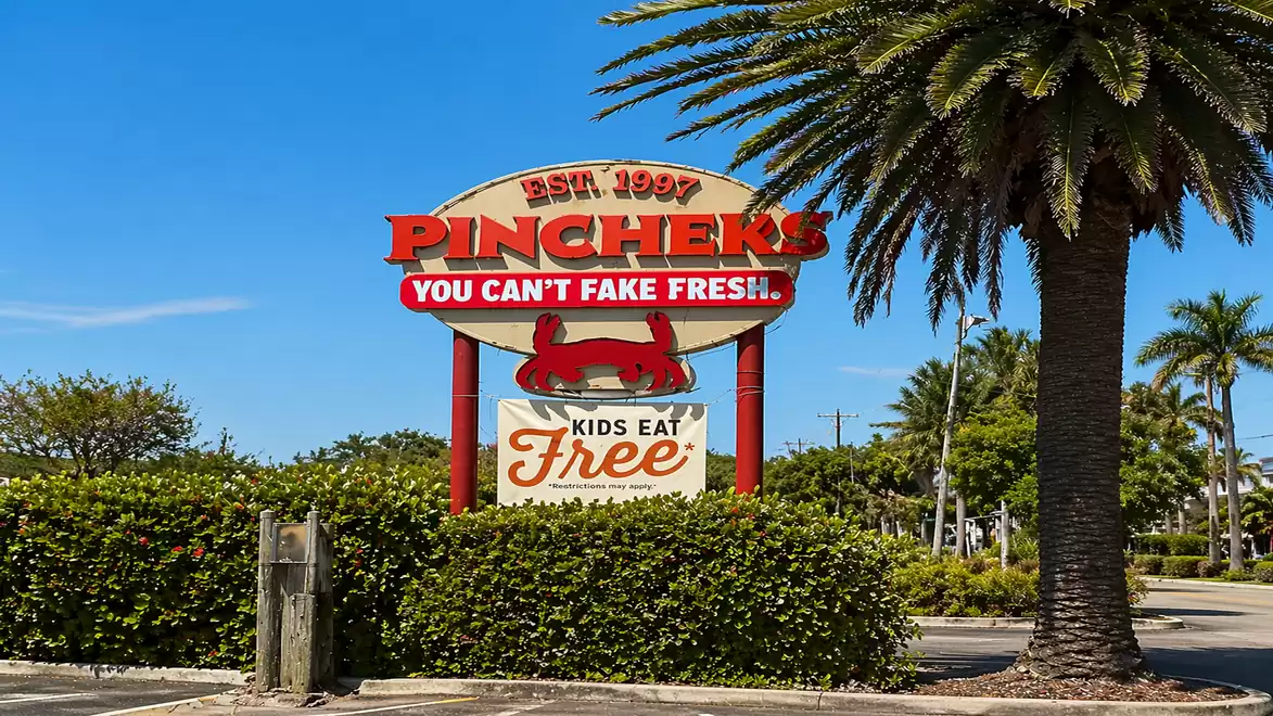 A sign for the restaurant Pinchers with the text 'You can't fake fresh' and 'Kids eat free'. Palm trees and blue sky in the background.