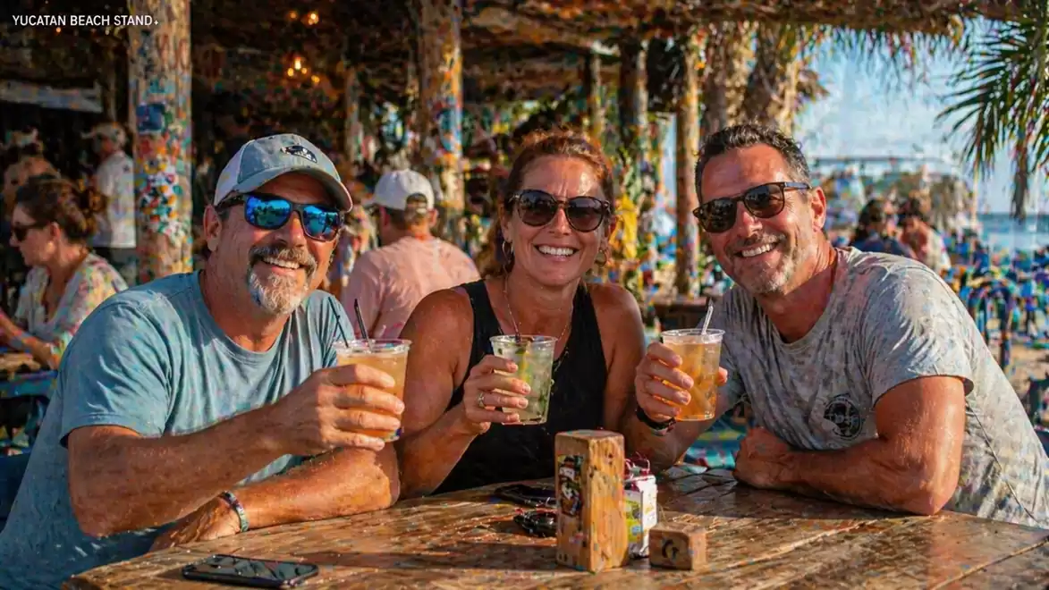 Three people sitting at an outdoor table holding drinks. They are dressed for warm weather and smiling at the camera. The setting is a beach bar or restaurant with decorative elements in the background.