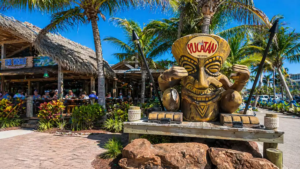 Large tiki statue in front of a tropical bar with a thatched roof, surrounded by palm trees under a bright blue sky.
