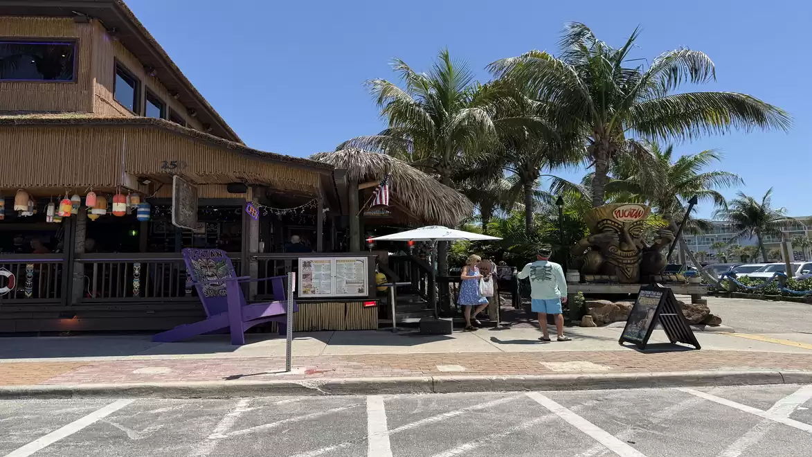 A tropical restaurant with a thatched roof, surrounded by palm trees. People are standing in front of the building, with a parking lot visible in the foreground.