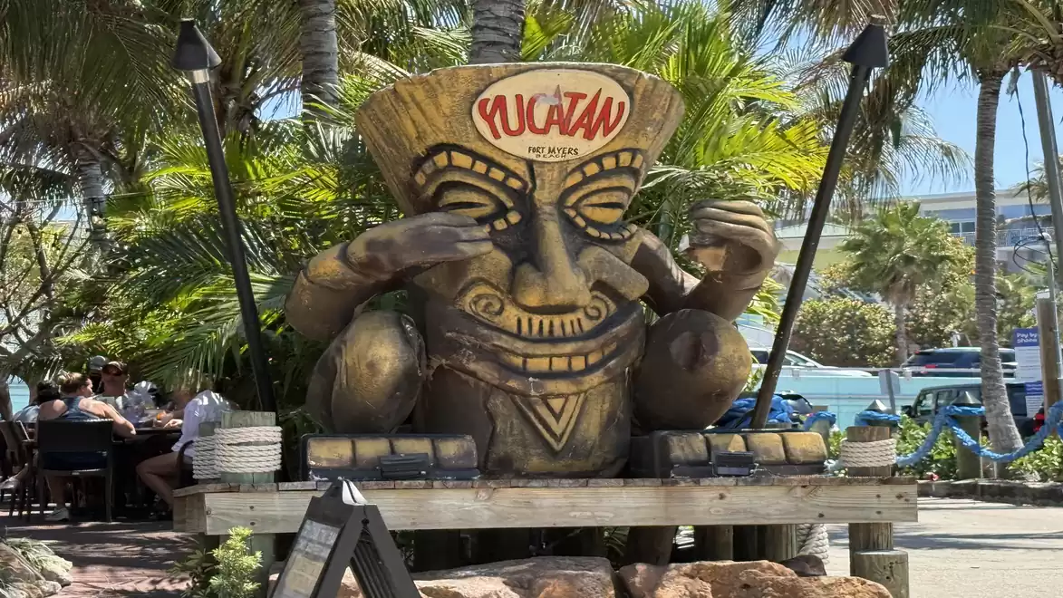 Jungle-like setting featuring a large wooden totem figure with the word 'Yucatan' on it. Figures are seated in front, surrounded by palm trees and lush vegetation.