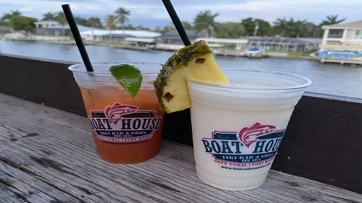 Two cocktails in plastic cups with boathouse logo, one garnished with pineapple and the other with a lime slice, on a wooden table by the water.