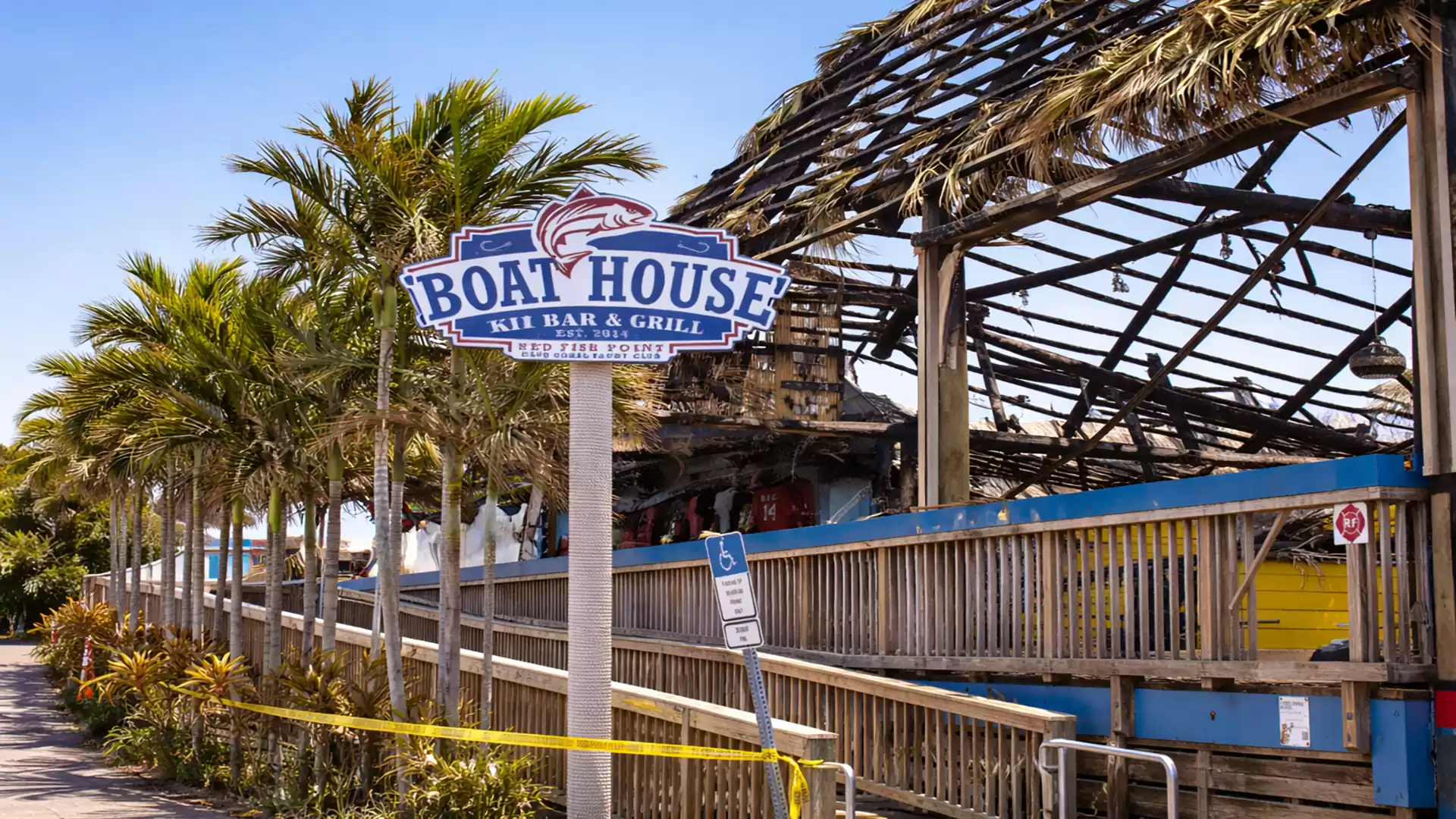 A damaged restaurant named 'Boat House Tiki Bar & Grill', surrounded by palm trees under a clear blue sky.