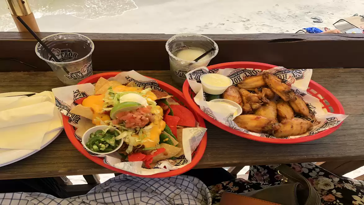 A table with a plate of nachos and chicken wings, served with sauce cups and two drinks in plastic cups. Sand is visible in the background.