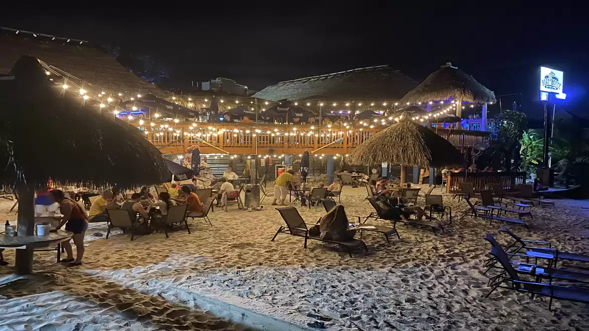 Night view of a lively beach with thatched huts and string lights casting a warm glow over the sand.