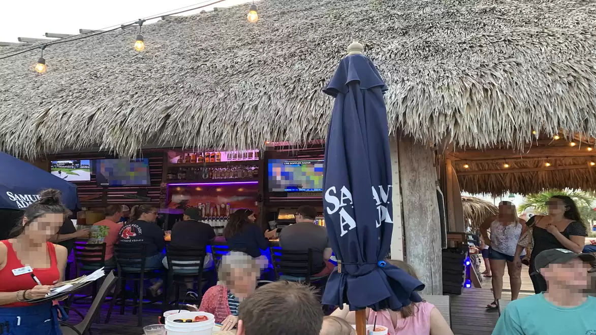 People sitting in a tiki bar with a thatched roof, enjoying drinks. TVs in the background are showing sports.