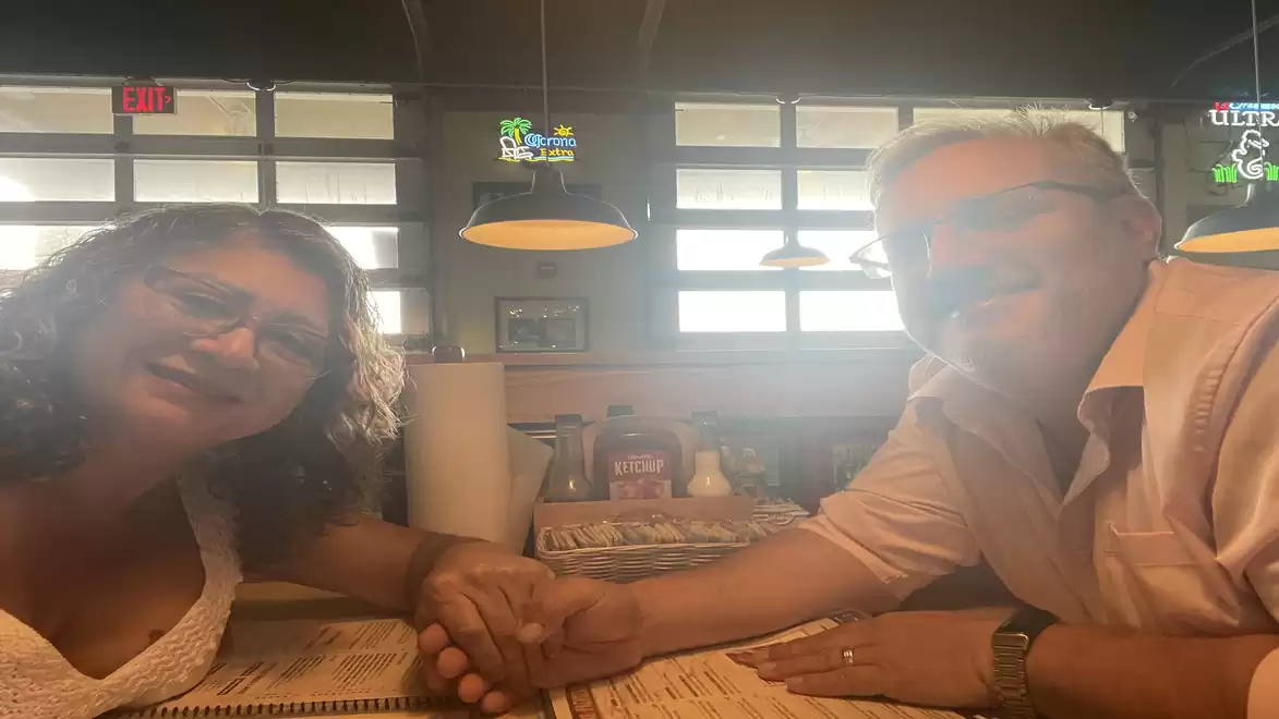 A couple is sitting in a restaurant holding hands. They appear happy and relaxed. Lamps and windows are visible in the background.