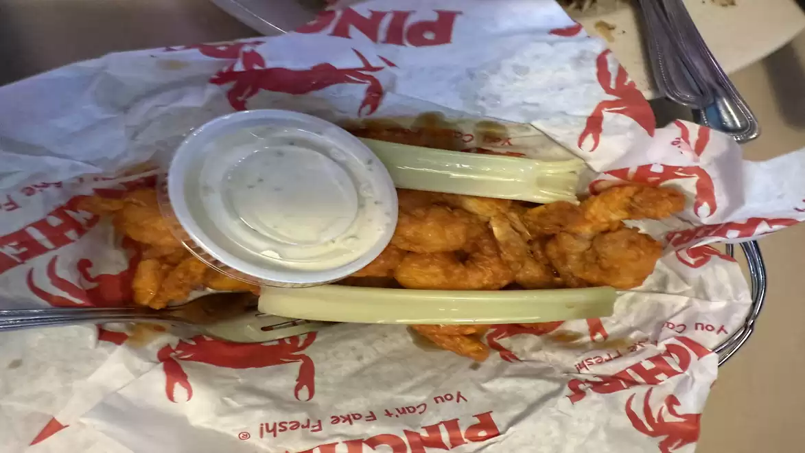 A plate of crispy fried shrimp, garnished with celery sticks and a cup of dip sauce, served on paper with a crab print.