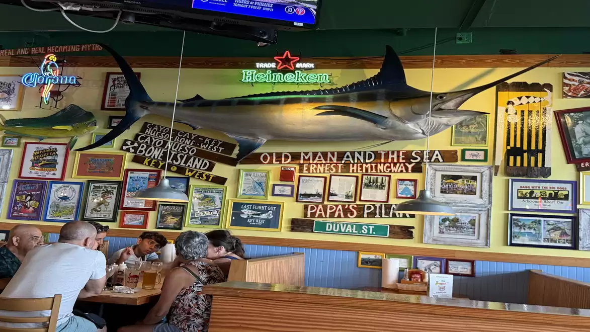 Interior shot of a restaurant with maritime decoration, including a large fish on the wall and various picture frames. People are sitting at a table conversing.