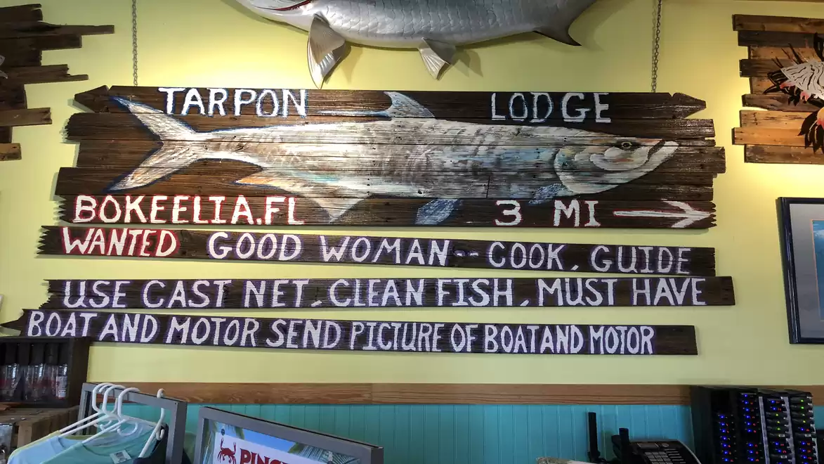 Wooden sign featuring a large fish painting and texts like 'Tarpon Lodge', 'Bokeelia FL 3 MI', and a humorous ad seeking a good woman and boat.