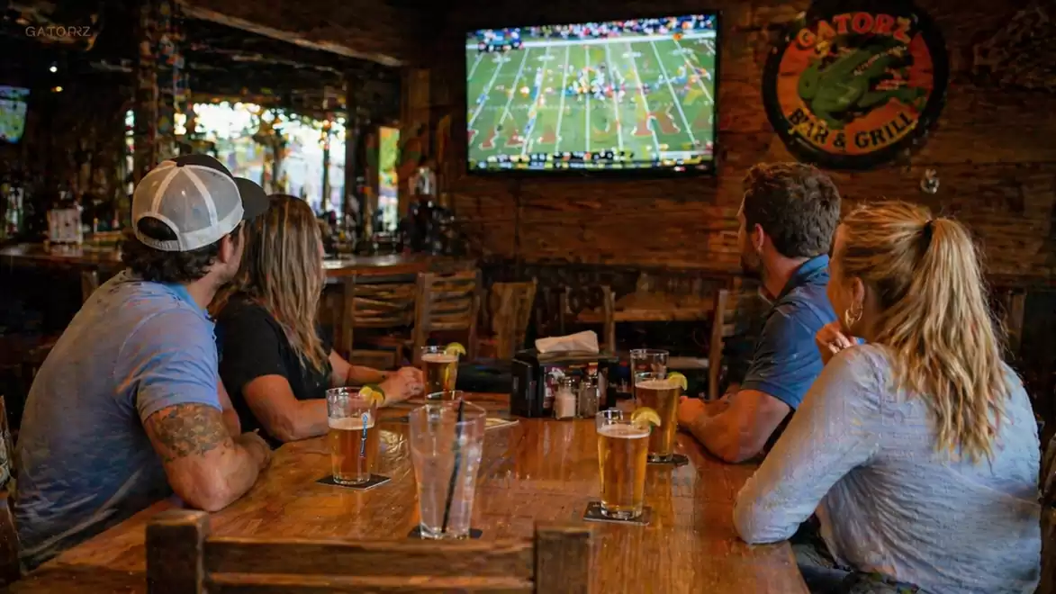 Four people watching a football game on TV in a bar while drinking beer.