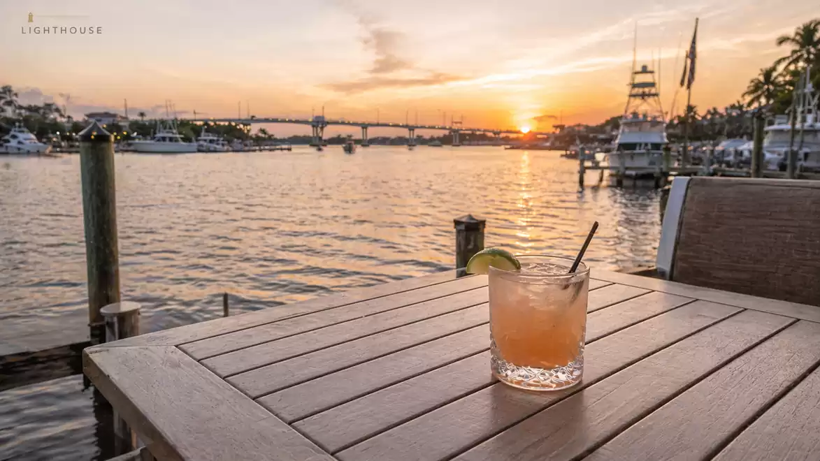 Wooden table with cocktail at sunset by a marina with boats and palm trees.
